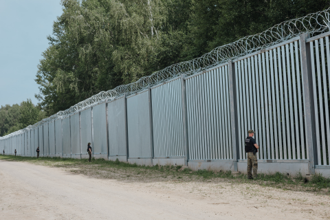 Stalowa, wysoka na kilka metrów zapora graniczna zakończona drutem kolczastym, przy której stoją strażnicy graniczni obserwujący między przęsłami przestrzeń za zaporą